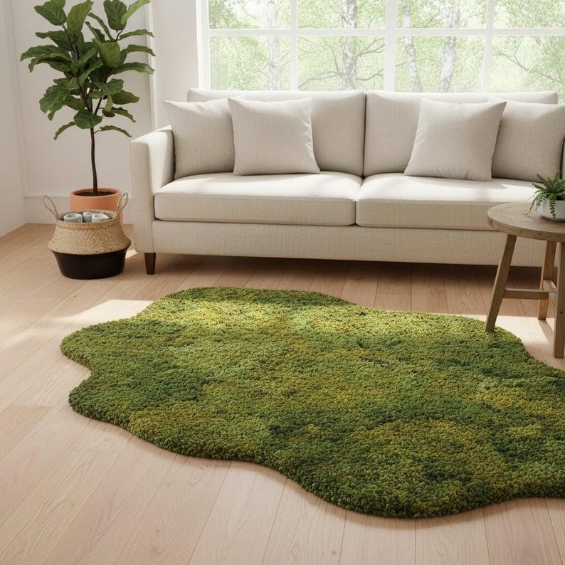 May include: A green, irregularly shaped rug with a moss-like texture. The rug is placed on a light wood floor in front of a cream-colored sofa. A potted plant and a small side table are also visible.