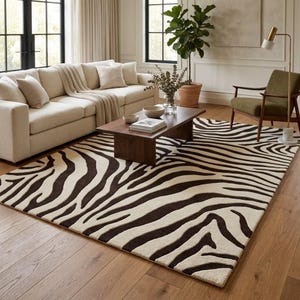 May include: A rectangular area rug featuring a high-contrast zebra stripe pattern in dark brown and cream. The rug is positioned on a light-toned wooden floor, covering a large portion of the space beneath a wooden coffee table and a beige sofa.
