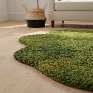 May include: A green, irregularly shaped rug with a textured, moss-like appearance. The rug's color transitions from lighter to darker shades of green, creating a natural, organic aesthetic. The rug is placed on a light-colored wooden floor.