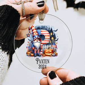 May include: A round glass ornament with a gnome wearing a red, white, and blue hat and outfit. The gnome is standing in front of a large letter "P" made of an American flag. The ornament also features a pumpkin, a haunted house, and the text "Paxten 2024".