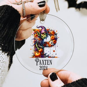 May include: A round, clear glass ornament with a green goblin character holding a pumpkin. The goblin is surrounded by colorful flowers and the ornament is personalized with the name "Paxten" and the year "2024".