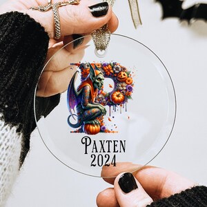 May include: A clear glass ornament with a green and purple gargoyle holding a pumpkin. The ornament is decorated with flowers and pumpkins. The text "PAXTEN 2024" is printed on the ornament.