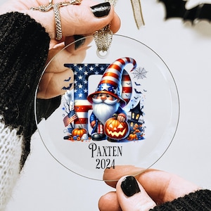 May include: A clear glass ornament featuring a gnome wearing a red, white, and blue hat and holding a pumpkin. The gnome is standing in front of a red, white, and blue American flag. The ornament also includes the text "Paxten 2024".