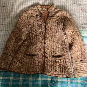 Puede incluir: Una chaqueta acolchada con estampado paisley en tonos marrones y rojos. La chaqueta tiene cuello mandarín, cierre de botones y dos bolsillos.