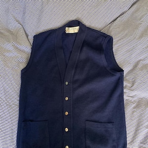 Può includere: Un cardigan blu navy senza maniche con scollo a V, cinque bottoni e due tasche. L'etichetta riporta "PURE WOOL / SCOTLAND".