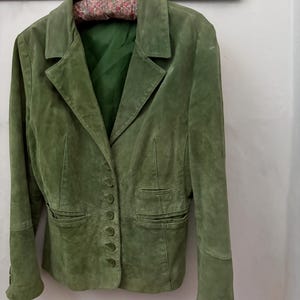 May include: A green suede blazer with a classic tailored design. The jacket features a notched collar, button closure, and two front pockets. The blazer is hanging on a floral print padded hanger.