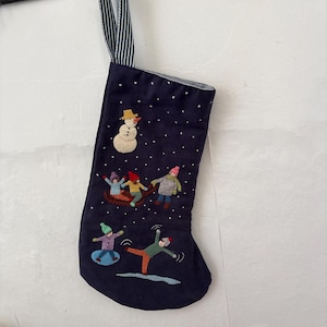May include: A dark navy blue Christmas stocking featuring embroidered winter scenes of a snowman and children playing in the snow. The stocking has a blue and white striped fabric hanging loop and is decorated with small white dots representing falling snow.