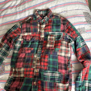 Può includere: Camicia a maniche lunghe con design patchwork. La camicia presenta un mix di motivi a quadri rossi, verdi, blu navy e crema. Ha due tasche sul petto e un colletto button-down. La camicia è realizzata in un materiale morbido, probabilmente cotone.
