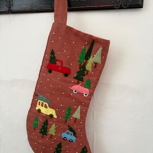 May include: A terracotta-coloured Christmas stocking featuring embroidered felt appliques of various cars in red, pink, yellow, and blue, each carrying a green pine tree on its roof, interspersed with small green pine trees and white embroidered snow dots.