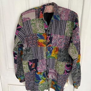 May include: A long-sleeved patchwork shirt with a button-down front. The shirt features a variety of colorful fabric patterns, including paisley, floral, and geometric designs in shades of green, purple, blue, and yellow. The shirt is hanging on a metal hanger.