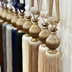 May include: A set of decorative tassel tiebacks in various colors, including brown, black, white, beige, and gold. The tassels have a gold metal finial and are made of a soft, silky material.