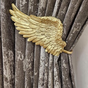 Puede incluir: Sujeta cortinas de metal dorado en forma de ala de ángel. El sujeta cortinas está sujetando un par de cortinas marrones con estampado.