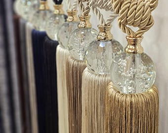 May include: Set of five curtain tiebacks with crystal finials and tassels. The tiebacks are made of braided rope in beige, cream, and brown colors.