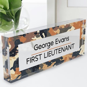 May include: A clear acrylic desk nameplate with a camouflage pattern in shades of brown, tan, and black. The nameplate has a white rectangle with the text "George Evans" and "FIRST LIEUTENANT" in black.