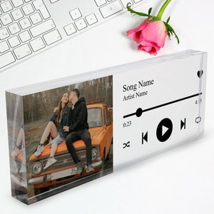 Pode incluir: Um bloco de acrílico transparente com uma foto de um casal sentado no capô de um carro laranja vintage. O bloco tem uma interface de reprodutor de música com o texto "Song Name" e "Artist Name" e botões de reprodução, pausa e avanço.