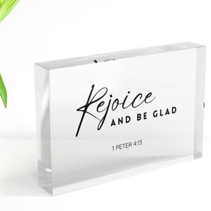 Può includere: Un blocco di acrilico trasparente con le parole "Rejoice and Be Glad" in corsivo nero. Il testo "1 Peter 4:13" è in basso. Il blocco rettangolare è su una superficie bianca.