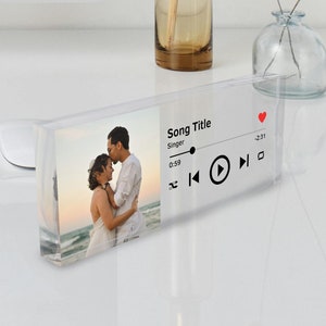 Pode incluir: Um bloco de acrílico transparente com uma foto de um casal na praia. O bloco também tem uma interface de reprodutor de música com o título da música "Song Title", o artista "Singer" e um botão de reprodução.