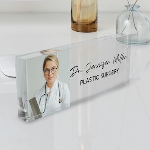 Pode incluir: Uma placa de nome de mesa de acrílico transparente com uma foto de uma mulher vestindo um jaleco branco de médico e um estetoscópio. O texto "Dr. Jennifer Miller PLASTIC SURGERY" está impresso na placa.