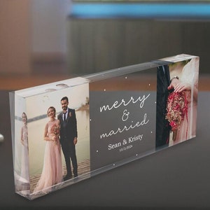 Pode incluir: Um bloco de acrílico transparente com duas fotos de um casal no dia do seu casamento. O texto "merry & married" está impresso no bloco, juntamente com os nomes do casal e a data do casamento.
