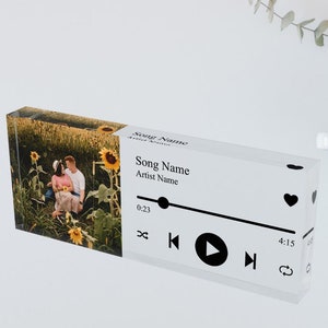 Pode incluir: Um leitor de música de acrílico transparente com uma foto de um casal em um campo de girassóis. O leitor tem um design preto e branco com um botão de reprodução, um botão de retrocesso, um botão de avanço rápido e um ícone de coração. O texto "Song Name" e "Artist Name" é exibido no leitor.