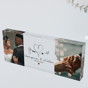 Pode incluir: Um bloco de acrílico transparente com duas fotos de um casal no dia do seu casamento. A primeira foto mostra o casal se olhando, e a segunda foto mostra suas mãos com suas alianças de casamento. O texto "g & w Georgina & Wilton 08.24.2024" está impresso no bloco.