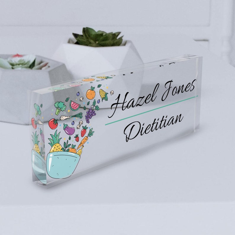 May include: A clear acrylic desk nameplate with a colorful illustration of a bowl of fruit and vegetables. The nameplate reads "Hazel Jones, Dietitian."