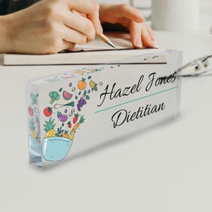 May include: A clear acrylic desk nameplate with a colorful illustration of a bowl of fruit and vegetables. The nameplate reads "Hazel Jones, Dietitian".