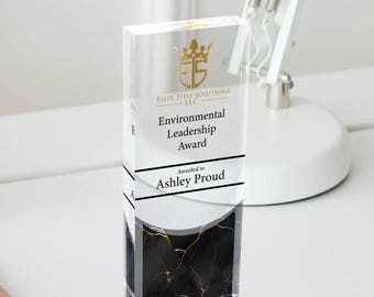 Puede incluir: Un premio de acrílico transparente con un diseño de mármol negro y dorado. El premio presenta el texto "Elite Title Solutions LLC", "Environmental Leadership Award" y "Awarded to Ashley Proud".