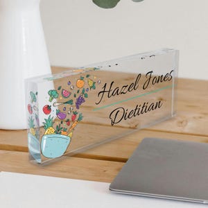 May include: A clear acrylic desk nameplate with a colorful illustration of a bowl overflowing with fruits and vegetables. The nameplate reads "Hazel Jones Dietitian".