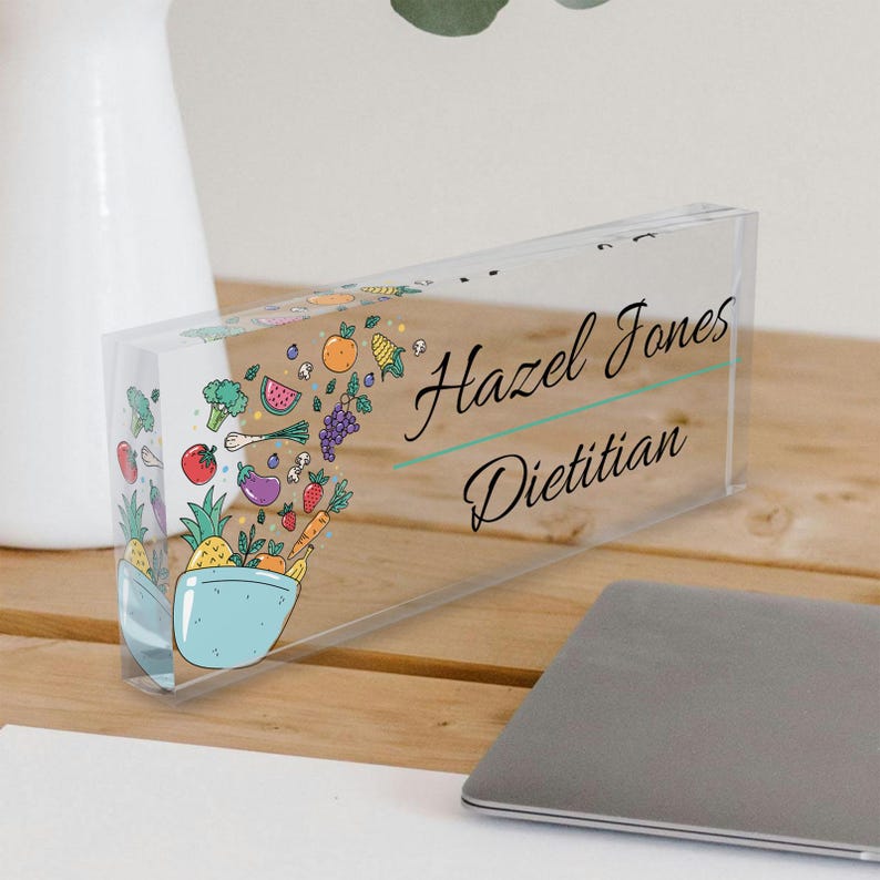 May include: A clear acrylic desk nameplate with a colorful illustration of a bowl overflowing with fruits and vegetables. The nameplate reads "Hazel Jones Dietitian".