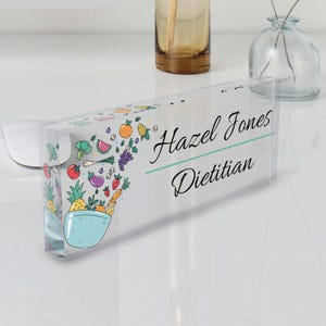 May include: A clear acrylic desk nameplate with a colorful illustration of a bowl overflowing with fruits and vegetables. The text "Hazel Jones Dietitian" is printed in black cursive font.