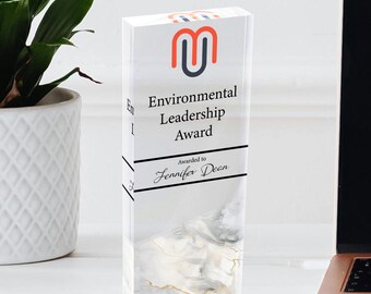 May include: A clear acrylic award with a marble design. The award features a red and gray logo with the letter "M" and the text "Environmental Leadership Award". The award is inscribed with the text "Awarded to Jennifer Dean".