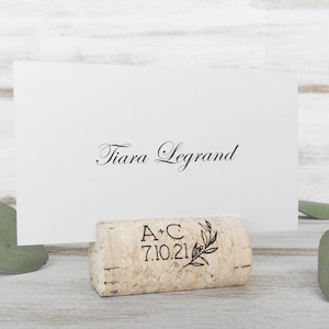 May include: Cork place card holder with a white card that says "Tiara Legrand". The cork is engraved with "A+C 7.10.21" and a small olive branch.