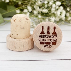 May include: Two wooden bottle stoppers. One is a cork stopper with a wooden base. The other is a wooden disc with the text "ANDERSON'S STOCK THE BAR" and the date "9.15.25" with three bottle illustrations.