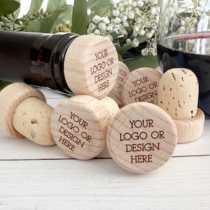 May include: A collection of bottle stoppers featuring natural cork bases and light-colored wooden tops. Each wooden top is engraved with the text 'YOUR LOGO OR DESIGN HERE'. The stoppers are arranged in a group, with one inserted into a dark glass bottle.