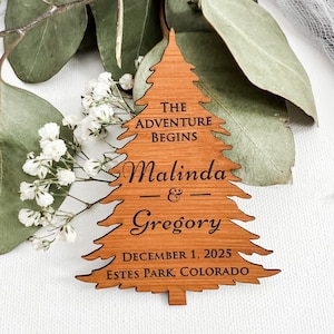 May include: A wooden Christmas tree ornament with the text "THE ADVENTURE BEGINS" and the names "Malinda & Gregory" along with the date "DECEMBER 1, 2025" and location "ESTES PARK, COLORADO". The ornament is brown and has a tree shape.