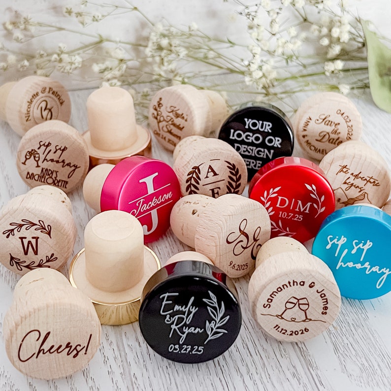 A collection of personalized wooden bottle stoppers with various designs and text. The stoppers are in different colors, including black, red, pink, blue, and brown. Some of the text on the stoppers includes 'Sip Sip Hooray',  'Save the Date', 'The Adventure Begins', and various names and wedding dates. The stoppers are a great way to add a personal touch to any event.
