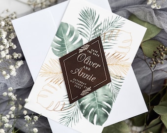 May include: Save the date card with a brown diamond-shaped tag with gold lettering that says "Save the Date Oliver and Annie Saturday October 25 2023". The card has a tropical theme with green and gold leaves.