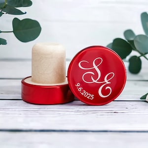 May include: A red bottle stopper with a wooden cork. The stopper's top features the white initials "S E" and the date "9.6.2025". The stopper is designed for wine bottles and is a decorative item.