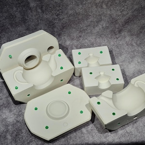 May include: A collection of white ceramic molds for making teapots and lids. The molds are in several pieces, showing the negative space of the teapot design. Green dots are visible on the molds, likely for alignment. The background is a textured gray surface.