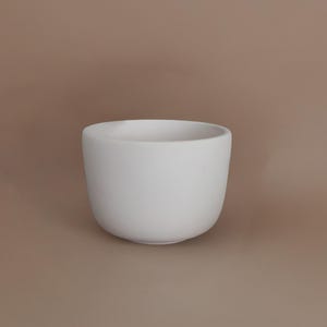 Puede incluir: Un cuenco de cerámica blanco con forma redondeada. El cuenco tiene un acabado liso y mate y una amplia abertura. El fondo es de color marrón neutro. Este cuenco es adecuado para servir alimentos o como objeto decorativo.