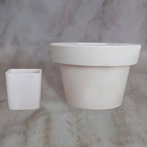May include: Two white ceramic plant pots. One is a small, square pot, and the other is a larger, round pot with a wide rim. The pots are empty and sit on a light-coloured surface.