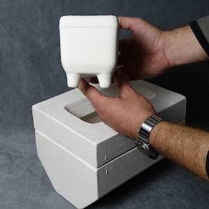 May include: A white, square-shaped ceramic container with three small legs is held above a matching rectangular base. The container has a lid and is being held by a person. The background is a solid gray color.