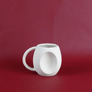 May include: A white ceramic mug with a distinctive, rounded shape and a large handle. The mug has a circular indentation on its side. It is set against a solid red background, emphasising its form and design.