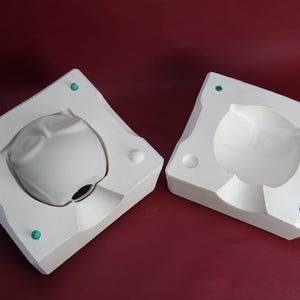 Pode incluir: Molde de coruja de cerâmica branca de duas partes para artesanato. O molde é em duas peças, com uma forma de coruja acabada em uma metade. O molde está sobre uma superfície bordô. O molde tem formato quadrado com bordas arredondadas.