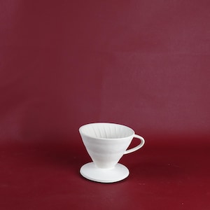 Peut inclure: Un filtre à café verseur en céramique blanche avec une poignée et une base circulaire. Le filtre à café est en forme de cône et repose sur un fond rouge uni. Le design est minimaliste et moderne.