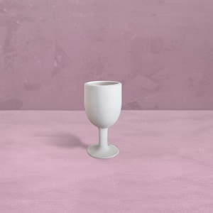 Könnte beinhalten: Ein kleiner, weißer Keramikbecher mit rundem Kelch und schlankem Stiel. Der Becher steht vor einem rosa Hintergrund. Der Becher wird wahrscheinlich zum Servieren von Getränken verwendet.