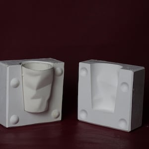 Può includere: Stampo in gesso bianco in due parti per creare una tazza o un contenitore di forma geometrica. Lo stampo è rettangolare con dettagli arrotondati e una cavità a forma di tazza sfaccettata. Sfondo bordeaux scuro.