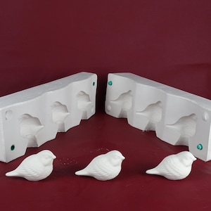 Puede incluir: Moldes de cerámica blanca para pájaros y tres pájaros de cerámica blanca acabados se muestran sobre una superficie burdeos. Los moldes son rectangulares con tres cavidades en forma de pájaro. Los pájaros acabados son pequeños y detallados.