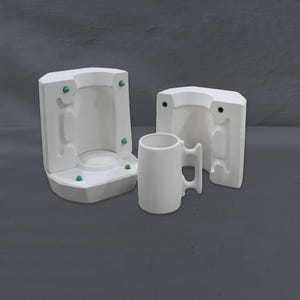 Puede incluir: Una taza de cerámica blanca con asa se encuentra junto a dos mitades de molde de cerámica blanca. Los moldes tienen cierres verdes. La taza es cilíndrica con un asa lateral. El fondo es gris liso.